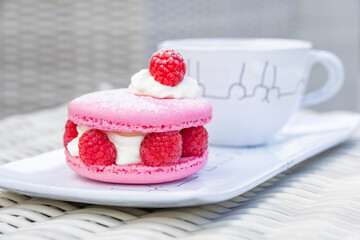 Elegant pink raspberry macaron with cream filling and fresh berries served alongside coffee on white porcelain plate. Perfect French pastry dessert presentation.
