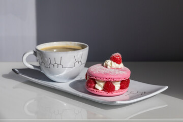 Elegant pink raspberry macaron with cream filling and fresh berries served alongside coffee on white porcelain plate. Perfect French pastry dessert presentation.