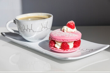 Elegant pink raspberry macaron with cream filling and fresh berries served alongside coffee on white porcelain plate. Perfect French pastry dessert presentation.