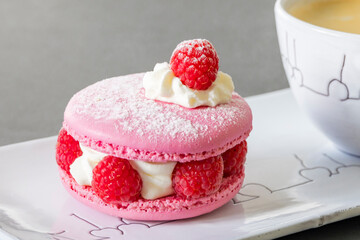 Elegant pink raspberry macaron with cream filling and fresh berries served alongside coffee on white porcelain plate. Perfect French pastry dessert presentation.