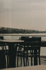 Outdoor dining area with a view of the lake on an overcast day. A dark table, chairs are arranged on a deck, with a serene lake view visible through the glass railing, creating a tranquil atmosphere.