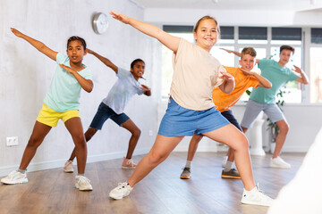Fototapeta premium Smiling cheerful tween girl practicing modern dances with group of children in studio, doing dabbing move..