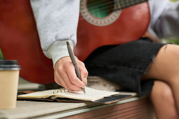 Caucasian young adult woman sitting outdoors holding acoustic guitar while writing in notebook with pen, takeaway coffee cup on bench, focusing on creative process