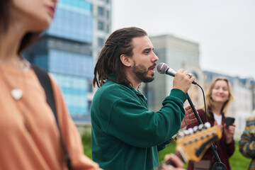 Caucasian young adult man singing into microphone outdoors with group of people playing instruments and smiling, diverse friends enjoying live music performance in urban setting