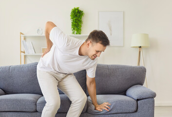 Sick man with lower back pain standing up from sofa in living room at home, holding lumbar with...