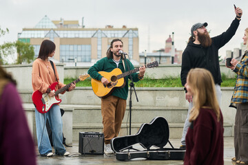 Multiethnic group of young adults performing live music outdoors, Caucasian man singing and playing acoustic guitar, Asian woman playing electric guitar