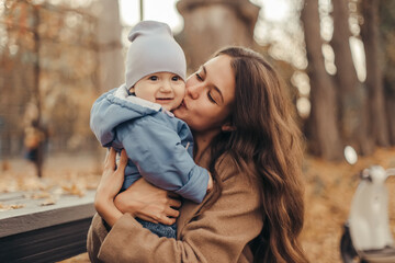 Fototapeta premium Young and happy mom dressed in cozy knitted clothes walking and having fun with her little son in the autumn park. Happy woman playing with her child outdoors.