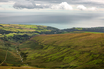 Fototapeta premium Laxey, Isle of Man 08,05,2023 Beautiful green hillside with a river running through it. The sky is cloudy and the ocean is in the background Isle of Man -