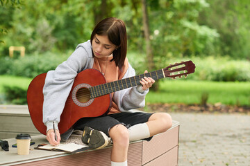Teenage Caucasian girl sitting outdoors playing acoustic guitar while writing notes in notebook, focusing on music composition, casual clothing, takeaway coffee cup on bench nearby