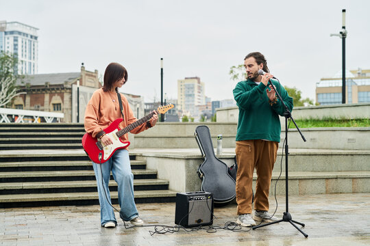 Asian young adult woman playing electric guitar while Caucasian young adult man adjusting microphone outdoors on city street, both standing near amplifier and guitar case, performing live music - Powered by Adobe