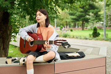 Teenage Caucasian girl sitting outdoors playing acoustic guitar and singing, wearing wireless earbuds, takeaway coffee cup and smartphone placed beside her on wooden bench in park
