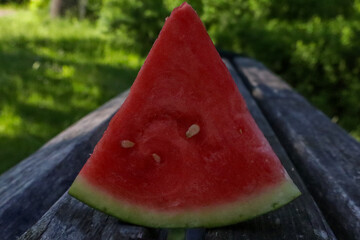 Close-up of a slice of ripe watermelon placed on a wooden surface, with bright red flesh, seeds visible, and surrounded by green nature in the background, perfect for summer refreshment.