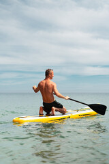 Back view of shirtless man paddling on kneeling position on paddleboard. Calm ocean and open sky create tranquil atmosphere of solo sporty retreat.