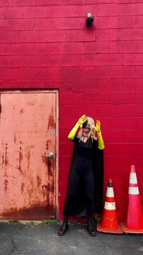 A woman is dancing outside with a cape and a mask against a city brick wall with a door and cones for a funny woman empowerment or confidence concept.