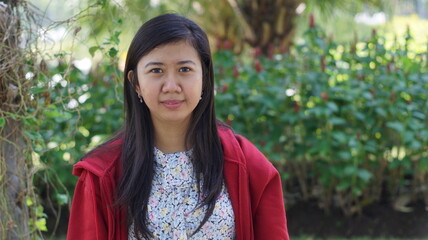 Portrait of a young Asian woman wearing a red jacket and floral blouse, standing outdoors with green plants and natural background, looking calm and confident