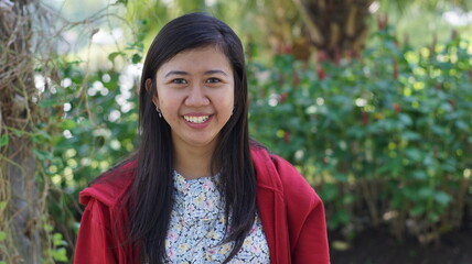 Portrait of a young Asian woman wearing a red jacket and floral blouse, standing outdoors with green plants and natural background, looking calm and confident