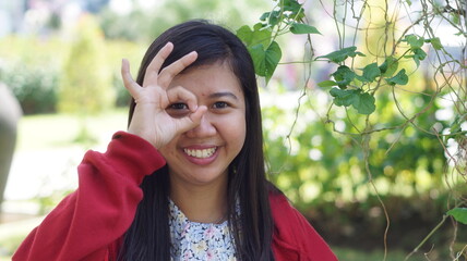 Portrait of a young Asian woman wearing a red jacket and floral blouse, standing outdoors with green plants and natural background, looking calm and confident