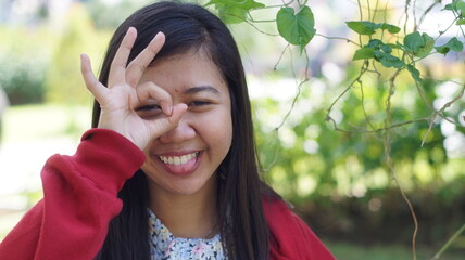 Portrait of a young Asian woman wearing a red jacket and floral blouse, standing outdoors with green plants and natural background, looking calm and confident