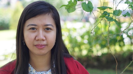 Portrait of a young Asian woman wearing a red jacket and floral blouse, standing outdoors with green plants and natural background, looking calm and confident