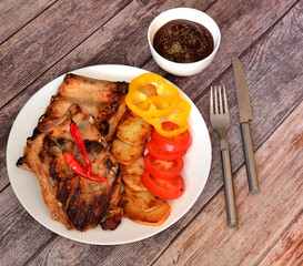 Fried pork ribs with potatoes, sweet and hot peppers, tomatoes, sauce and cutlery on a wooden table.