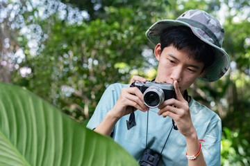 A young Asian man in a camouflage boonie hat holds a camera, taking a photo in a dense, green...