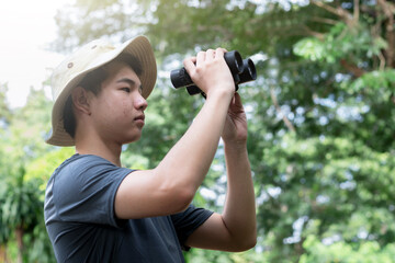 A young Asian man in a boonie hat is looking through binoculars and watching birds in a lush, green forest, enjoying nature and outdoor activities. He looks focused and ready for adventure.