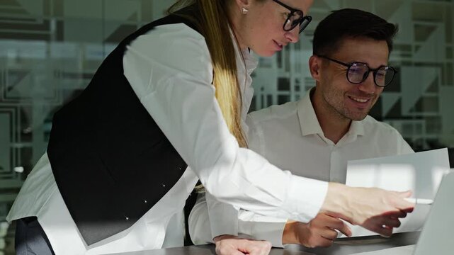 Two professionals are engaged in a collaborative effort in a modern office. One is pointing at the laptop screen, providing insights while the other takes notes, both focused on the task at hand.
