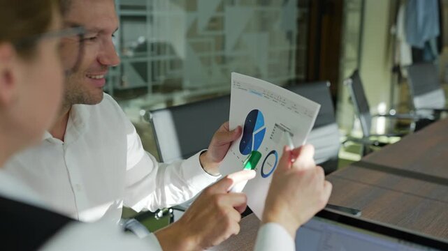 A man points to a chart while seated at a table with a woman. Both are engaged in discussion, using a tablet and paperwork to illustrate business metrics. They appear focused and professional.