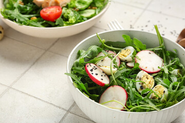 Bowl with tasty arugula salad on white tile background, closeup