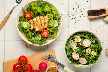 Bowls with tasty arugula salad and ingredients on white tile background