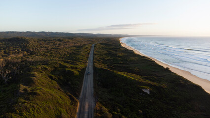 This aerial shot captures a straight, empty road running parallel to a long, sandy coastline. The road serves as a clear division between the dense, dark green forest and the wide, pale beach. 