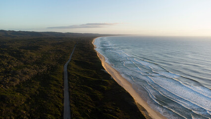 This aerial shot captures a straight, empty road running parallel to a long, sandy coastline. The road serves as a clear division between the dense, dark green forest and the wide, pale beach. 