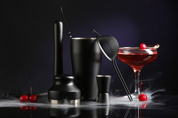 Bartender utensils, glass with cherry cocktail on table with smoke near black wall, closeup
