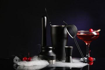 Bartender utensils, glass with cherry cocktail on table with smoke near black wall