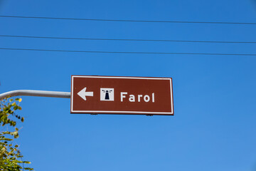 Fototapeta premium A sign where it reads in Portuguese the following text: Lighthouse. Brown road sign with arrow pointing left indicating direction to lighthouse.