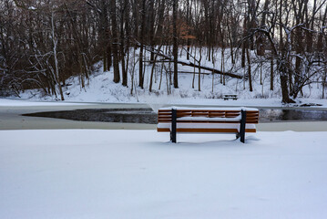 Snow covered bench overlooking partially frozen water