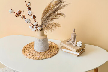 Vase with cotton flowers and pampas grass on table near beige wall in living room, closeup