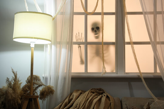 Interior of living room decorated for Halloween with window, skeleton and glowing lamp in evening