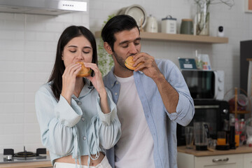 Happy couple eating burgers together in modern kitchen enjoying fast food meal symbolizing lifestyle choice