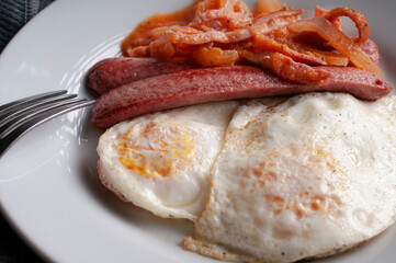 Macro photography of fried overeasy or over medium eggs with sausage, onion, and natural tomato sauce on a white plate, with a fork on the side, well done angle view