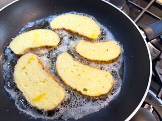Bread slices frying in oil on pan, bubbles around edges, gas stove in kitchen.