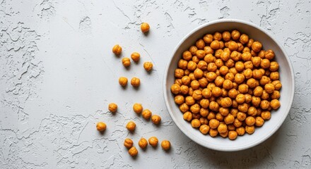 Fototapeta premium A bowl filled with roasted chickpeas, with some scattered on a textured light gray surface