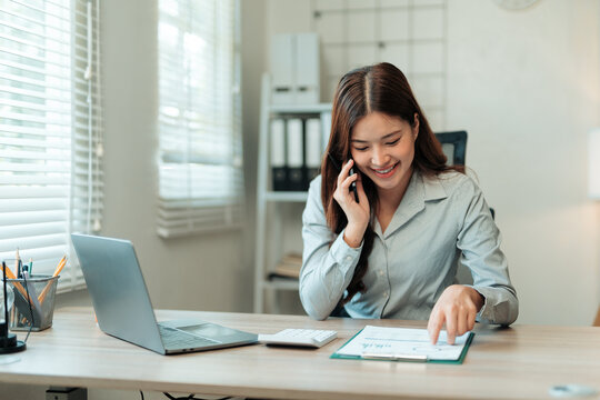 Smiling businesswoman analyzing financial charts while talking on mobile phone in modern office