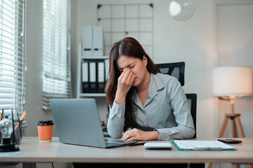 Young professional woman feeling tired and massaging her nose bridge while working on a laptop in the office