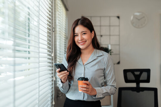 Young asian businesswoman holding mobile phone and drinking coffee standing near window in modern office - Powered by Adobe
