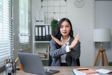 Young Asian businesswoman showing stop gesture with crossed arms, expressing denial, disagreement, or rejection in a modern office environment