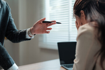 Business woman covering face with hands while manager is scolding her for bad work results pointing with pen sitting at office desk
