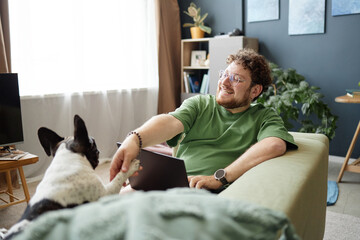 Caucasian young adult man sitting on sofa smiling and shaking paw with French Bulldog while working on laptop in living room, showing friendly interaction between pet and owner