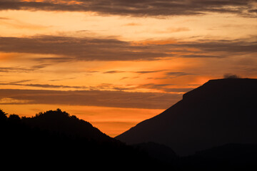 Beautiful silhouette of mountain peaks and forested hills during sunrise with dramatic orange and purple sky in the background.