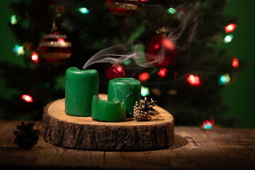 Green candles sit atop a wooden surface, emitting wisps of smoke. A beautifully decorated Christmas tree serves as a backdrop, adorned with vibrant lights and ornaments, creating a festive ambiance.